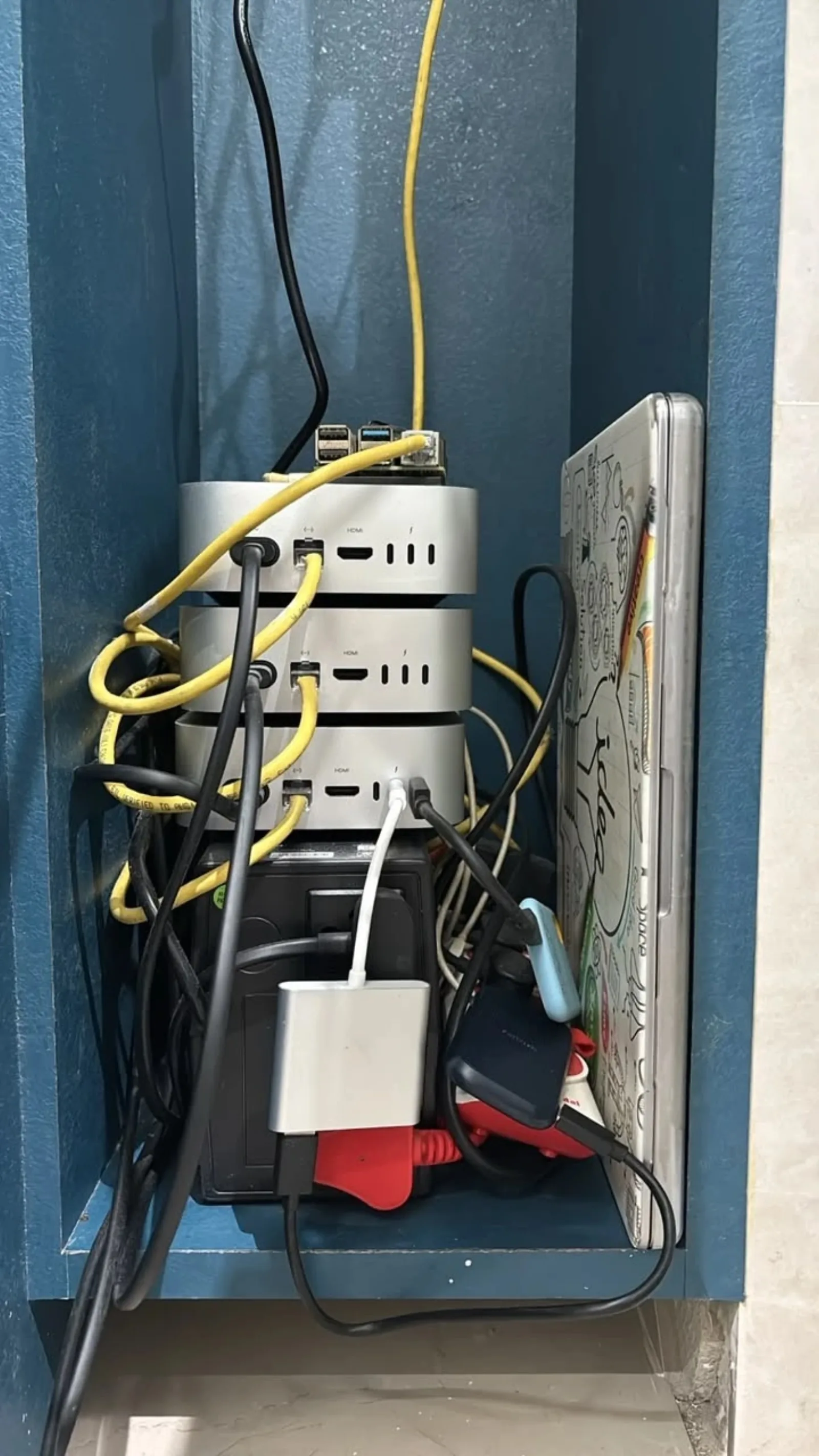 The cupboard at home: three Mac minis stacked with a Raspberry Pi on top, a UPS below and an old laptop beside them, cables everywhere