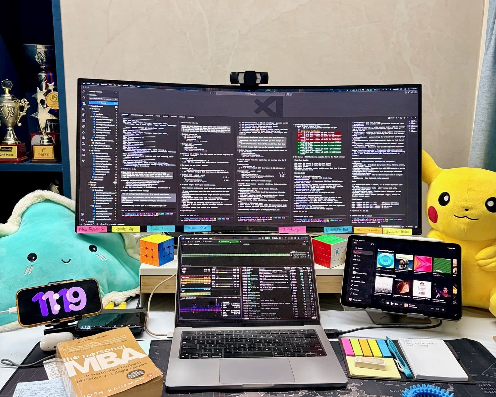 The desk — an LG ultrawide filled with agent panes, a MacBook Pro, an iPad as a second screen, sticky-note plans along the monitor's bottom edge, hackathon trophies on the left and a Pikachu on the right